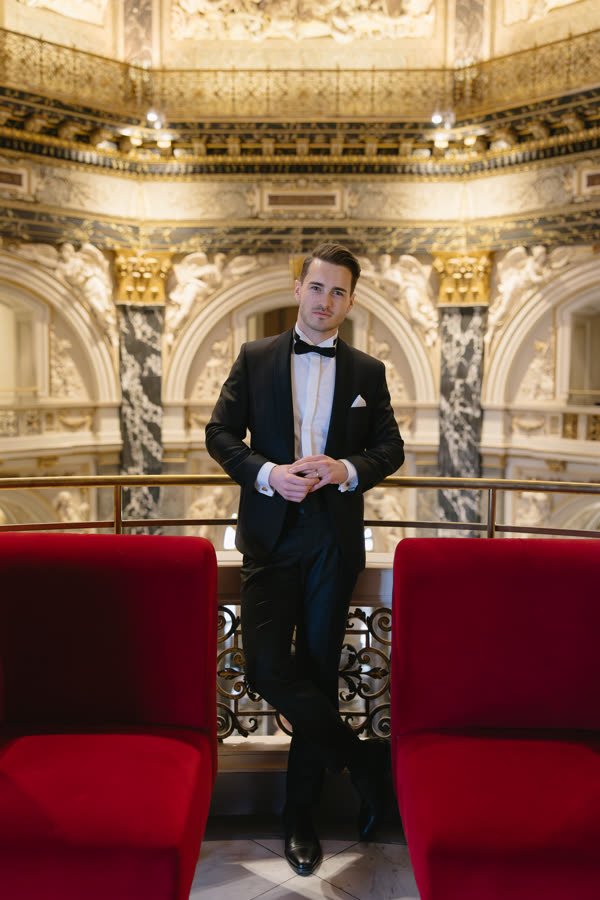 Andreas Schmid, tuxedo between red velvet chairs in a gilded museum hall